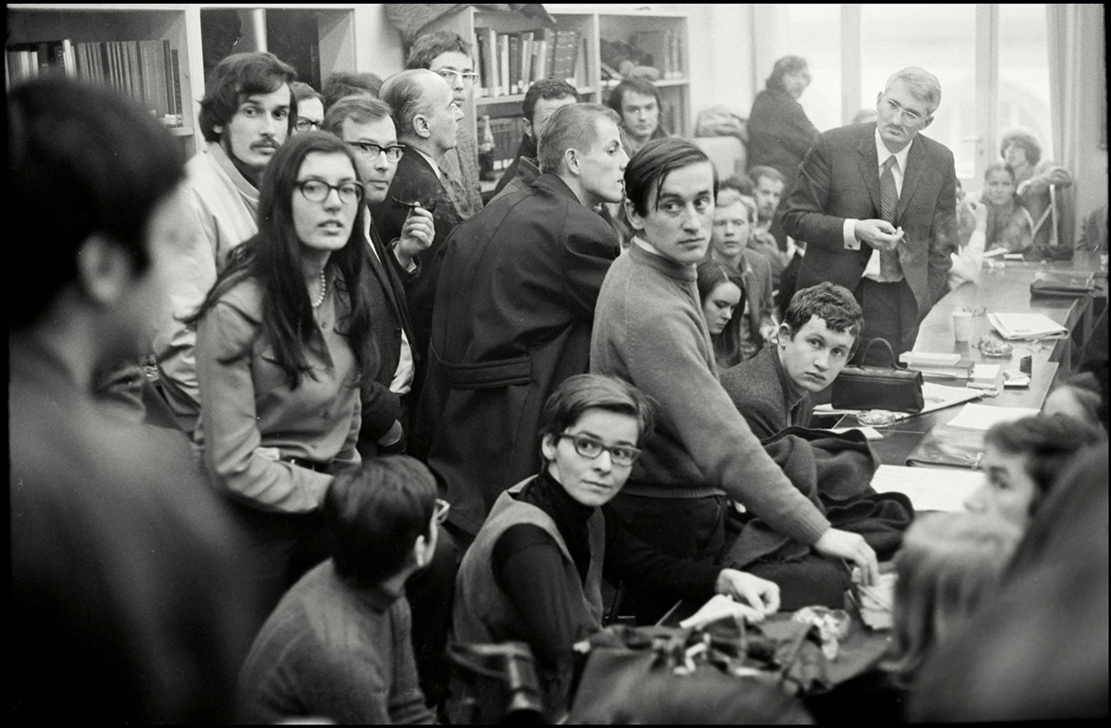 Black and white photo of a crowded room with people standing and sitting, some looking towards the camera, in a library or meeting.