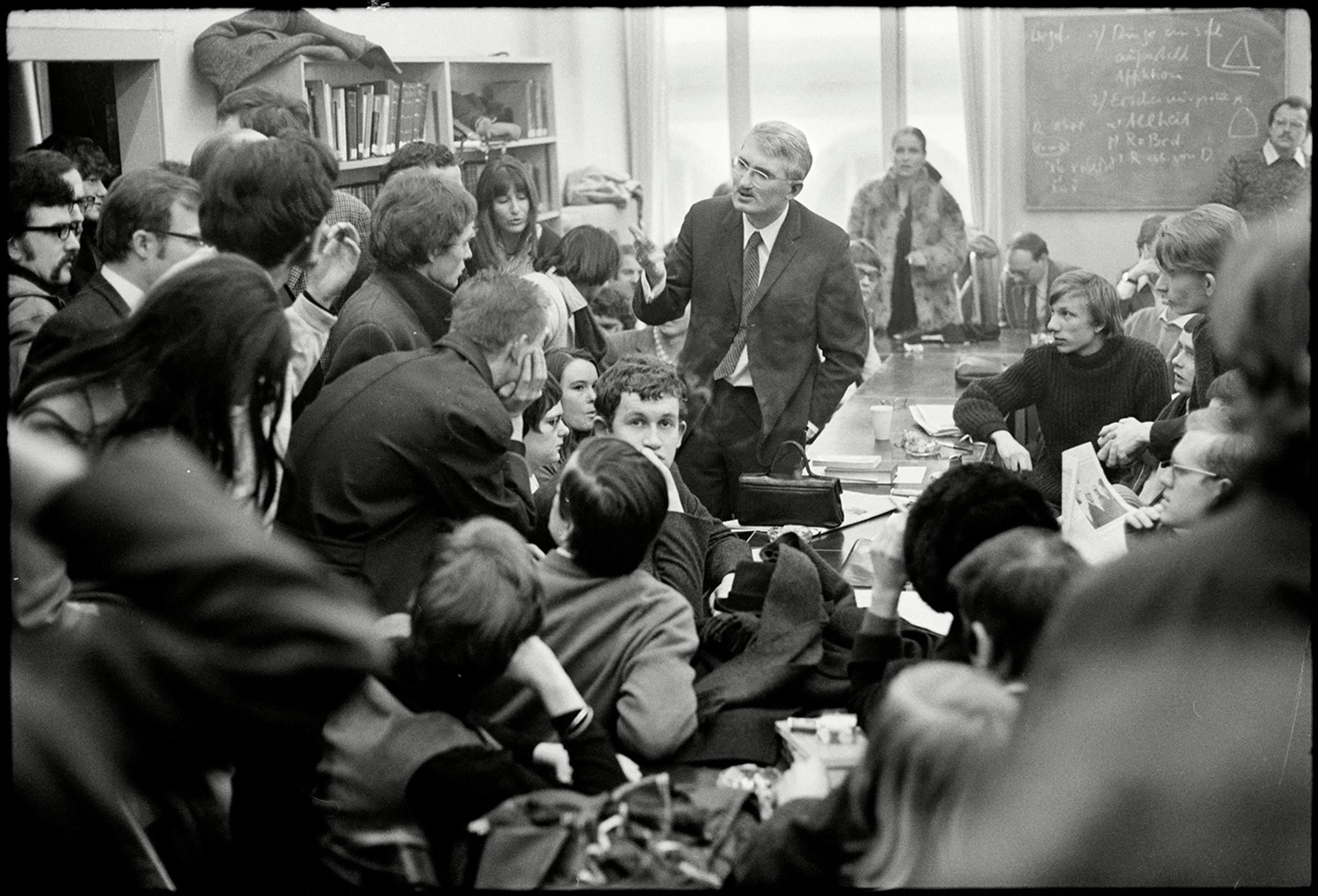 Black and white photo of a man speaking to a group of people in a crowded indoor setting with a blackboard in the background.