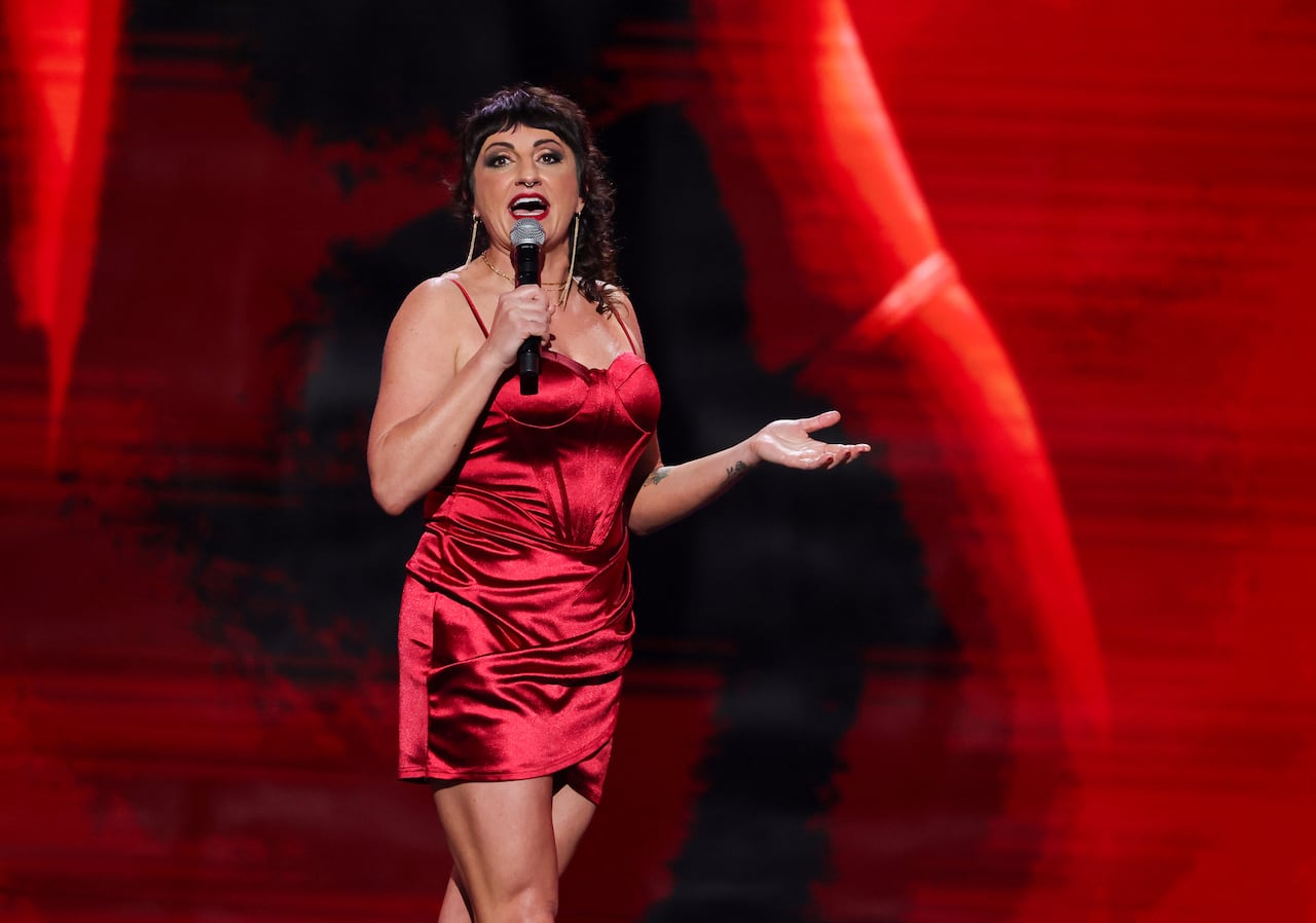 A woman with short brown bangs and a silky red corset dress stands on stage speaking into a mic