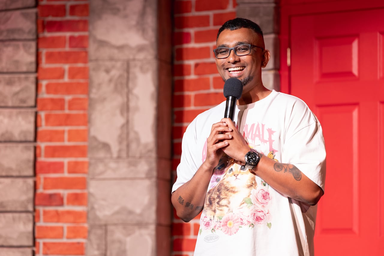 A smiling, bespectacled man in a kitten T-shirt speaks into a mic on stage against a backdrop of a brick wall and red door.