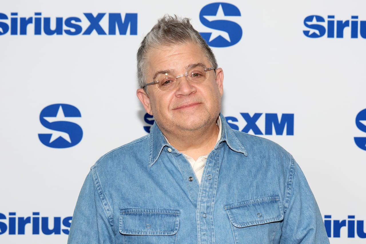 A man with graying hair, rimless glasses and a denim shirt stands in front of a backdrop with the words "SiriusXM" and an accompanying blue logo shaped like the letter S with a star in it.
