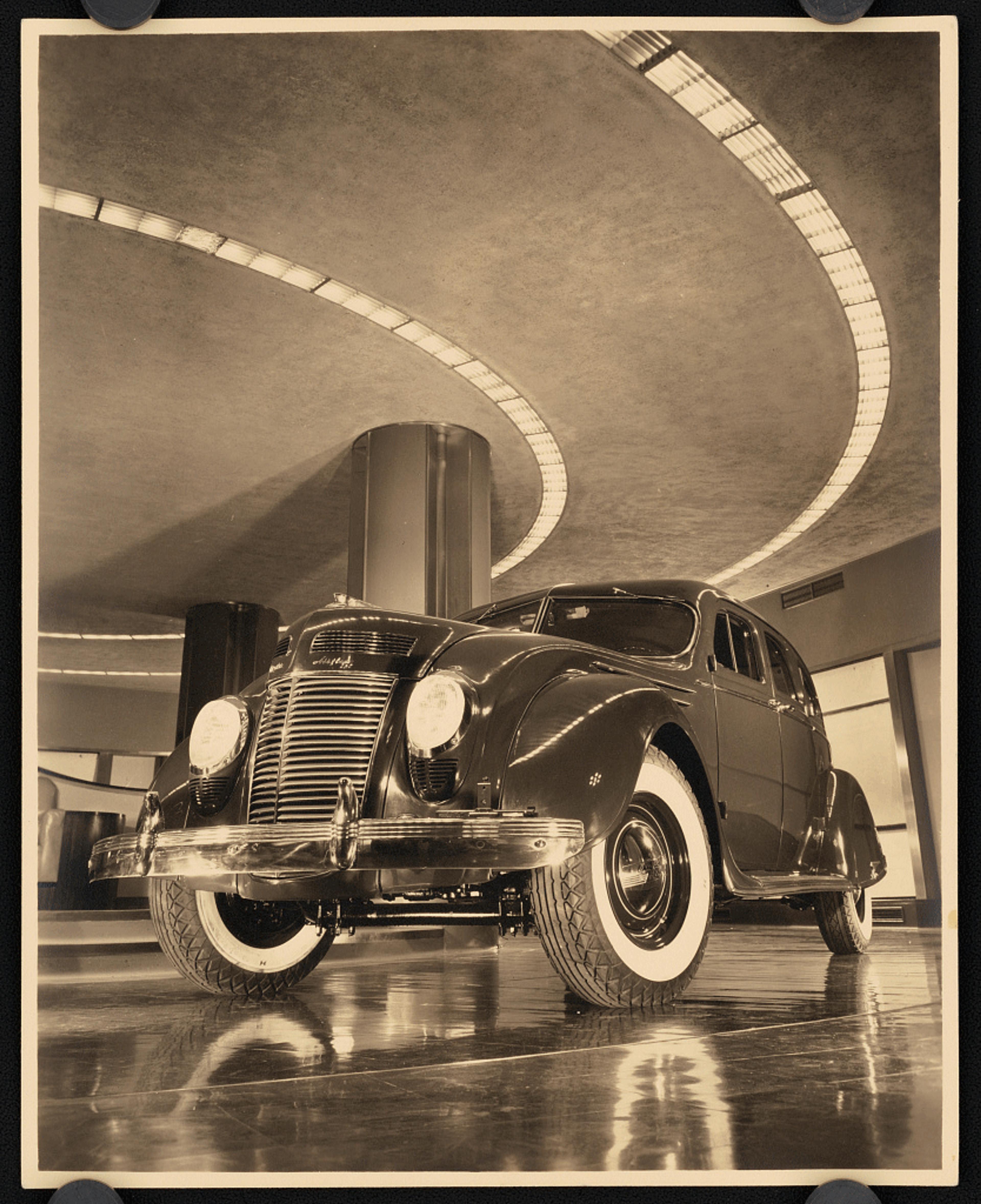 Sepia-toned photo of a vintage car on a shiny floor with curved ceiling lights above, creating a dramatic effect.