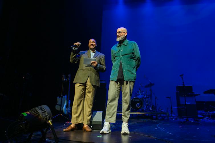 Two men stand on a stage and one holds a microphone.