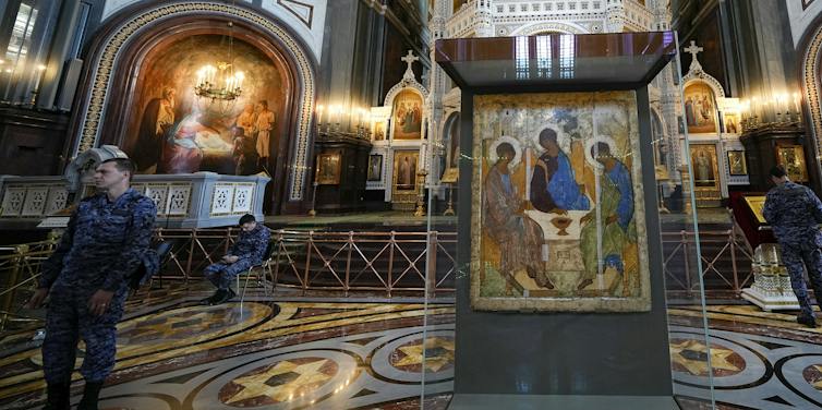Men in military fatigue stand next to a religious icon of three angel figures behind a class case inside an ornate church with gold decor.